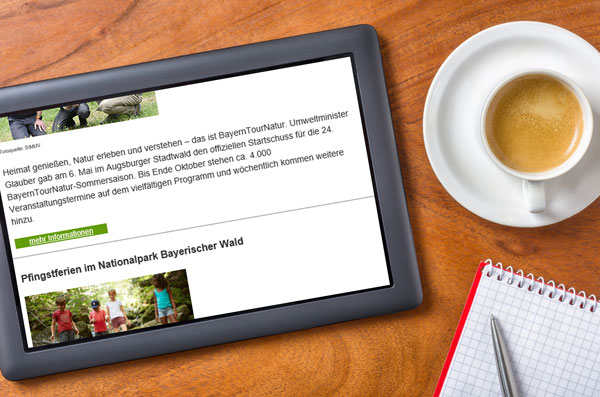 Tablet mit dem BayernTourNatur-Newsletter, daneben eine Tasse Kaffee sowie ein Block mit Stift